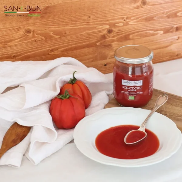 Na stole passata pomidorowa w talerzu i obok w szklanym słoiku. Obok talerza dwa pomidory w białym ręczniku.